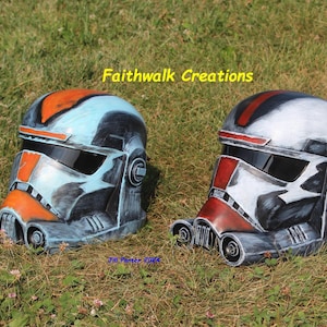 Könnte beinhalten: Zwei Helme im Science-Fiction-Stil mit dunklen Visieren. Der linke Helm hat eine verwitterte Lackierung in Hellblau, Orange und Schwarz. Der rechte Helm hat eine verwitterte Lackierung in Weiß, Rot und Schwarz. Text: Faithwalk Creations und Jill Panter 2024.