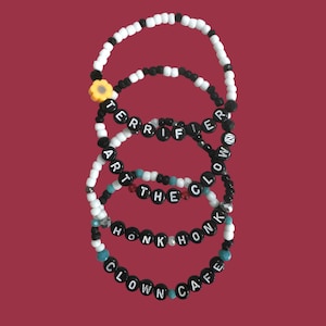 Op de afbeelding: Vier armbanden met zwarte en witte kralen, en tekst met de spelling "TERRIFIER", "ART THE CLOWN", "HONK HONK" en "CLOWN CAFE". Eén armband heeft een gele bloemkralen. De armbanden staan op een bordeauxrode achtergrond.