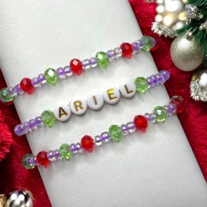 Könnte beinhalten: Drei Perlenarmbänder mit roten, grünen und violetten Perlen. Das mittlere Armband hat den Namen "ARIEL" in weißen Perlen mit goldenen Buchstaben.