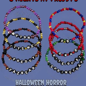 May include: Several beaded bracelets with text spelling out phrases like "Afton", "It's Me", and "Freddy Fazbear". The bracelets are in various colors, including red, purple, and black. The image also includes the text "5 Nights at Freddy's" and "Halloween Horror Nights 34".