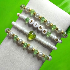 Puede incluir: Cuatro pulseras con cuentas blancas, cristales verdes y detalles dorados. Las pulseras deletrean la palabra "TINK" con cuentas de letras blancas. Una pulsera tiene un dije de corazón verde.