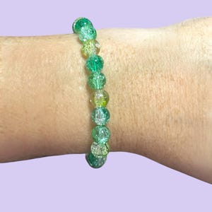 Puede incluir: Una pulsera de cuentas verdes con varios tonos de verde y cuentas transparentes. La pulsera se lleva en una muñeca sobre un fondo morado claro. Las cuentas tienen un aspecto agrietado.