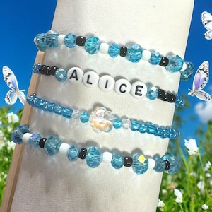 May include: Four beaded bracelets in shades of blue, white, and black. One bracelet spells out "ALICE" with white letter beads. The bracelets feature various bead shapes and sizes, with a butterfly and floral background.