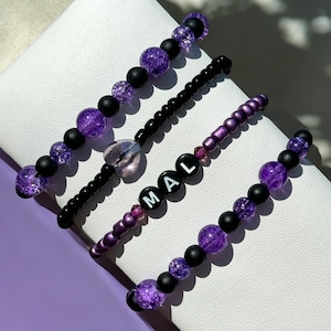 May include: Four bracelets with purple and black beads. One bracelet has the word "MAL" spelled out in black beads.