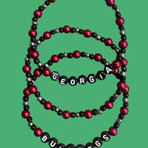 May include: Three beaded bracelets with a green background. The bracelets are made of red, black, and silver beads. One bracelet spells out "GEORGIA" and another spells out "BULLDOGS" using white letter beads.