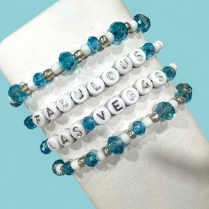 Peut inclure: Quatre bracelets de perles avec des lettres blanches épelant "FABULOUS VEGAS". Les bracelets présentent des perles turquoise et argentées. Les bracelets sont exposés sur une surface blanche sur fond bleu clair.