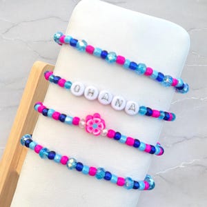 May include: Four beaded bracelets with blue, pink, and white beads. One bracelet has the word "OHANA" spelled out in white letters. Another bracelet has a pink flower charm.