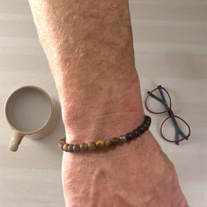 Könnte beinhalten: Ein Perlenarmband mit braunen, gelben und schwarzen Steinen wird am Handgelenk getragen. Eine beige Tasse und eine Brille mit roten und blauen Gläsern liegen auf einer hellen Oberfläche.