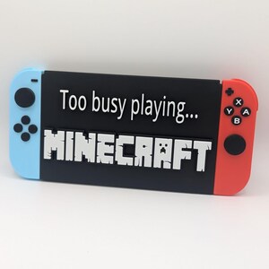 Puede incluir: Una consola Nintendo Switch negra con un mando azul y rojo. La consola tiene una pegatina blanca que dice "Demasiado ocupado jugando... MINECRAFT".