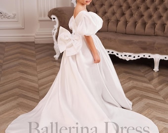 May include: A white satin ballerina dress with a large bow on the back and puffed sleeves. The dress has a long train and is from the Ballerina Dress Boutique. The girl is standing in front of a brown sofa.