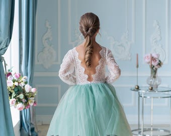 Può includere: Una bambina indossa un abito di tulle verde chiaro con un corpetto in pizzo bianco e maniche lunghe. L'abito ha una schiena scoperta e i capelli della bambina sono raccolti in una coda di cavallo. L'abito è di Ballerina Dress Boutique.