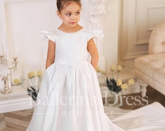 May include: A young girl wearing a white satin dress with a pearl trim. The dress has a full skirt and a train. The girl is standing in front of a white fireplace.