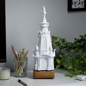 May include: A white 3D printed model of a building with a spire and a clock face. The model is on a wooden base and is sitting on a white desk with a potted plant, pencils, and a candle.