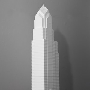 May include: A white architectural model of a skyscraper, featuring a tiered design and a pointed top. The model is placed on a wooden surface with a black metal frame, set against a gray backdrop. The building model is a detailed representation of a tall structure.