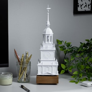 May include: White 3D printed model of a clock tower with a wooden base. The model is on a desk with a potted plant, a jar of pens, and a candle.