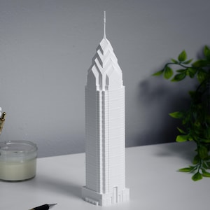 May include: A white, 3D-printed model of a skyscraper, featuring a detailed architectural design. The building has a spire at the top. The model is on a white surface, with pens and pencils in a jar to the left and greenery to the right.