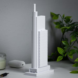 May include: A white paper model of a tall skyscraper with a modern design. The model is sitting on a white surface with a potted plant in the background.
