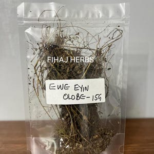 Può includere: Un sacchetto di plastica trasparente pieno di erbe essiccate. L'etichetta bianca riporta la scritta "EWE EYIN OLOBE-15G". Il marchio "FIHAJ HERBS" è stampato sul sacchetto. Le erbe sono marroni e verdi.