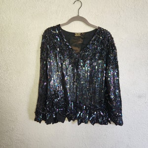 May include: A black sequined top with iridescent sequins. The top has a V-neck and long sleeves. The hemline is scalloped.