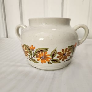 May include: A white ceramic jar with a speckled glaze and a floral design in shades of orange, brown, and green. The jar has two handles and a rounded body.
