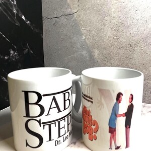 May include: Two white ceramic mugs. One mug has the text "BABY STEP" in large black letters, with "Dr. Leo Marvin" below. The other mug features a scene with two men and the text "What About Bob?"