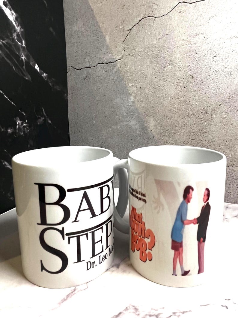 May include: Two white ceramic mugs. One mug has the text "BABY STEP" in large black letters, with "Dr. Leo Marvin" below. The other mug features a scene with two men and the text "What About Bob?"