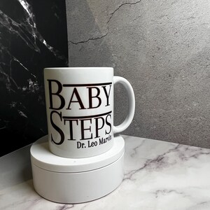 May include: White ceramic mug with the words "BABY STEPS" in black, with "Dr. Leo Marvin" below. The mug is on a white, round pedestal, set against a marble and grey background.