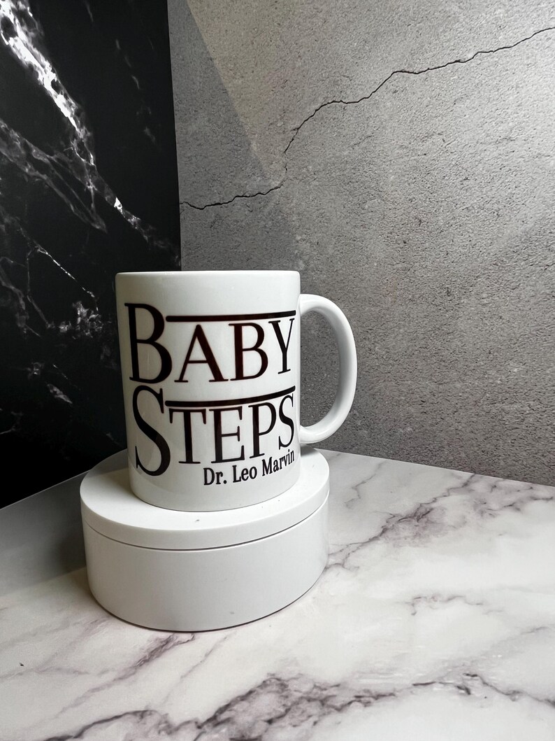 May include: White ceramic mug with the words "BABY STEPS" in black, with "Dr. Leo Marvin" below. The mug is on a white, round pedestal, set against a marble and grey background.