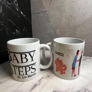 May include: Two white ceramic mugs. One mug has the text "BABY STEPS" in black, with "Dr. Leo Marvin" below. The other mug features the text "What about Bob?" in orange and a graphic of two people.