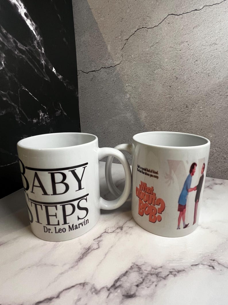 May include: Two white ceramic mugs. One mug has the text "BABY STEPS" in black, with "Dr. Leo Marvin" below. The other mug features the text "What about Bob?" in orange and a graphic of two people.