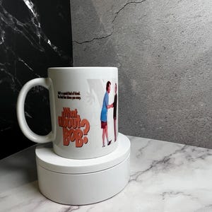 May include: White ceramic mug featuring the text "What About Bob?" in orange and an image of two men shaking hands. The mug also has the quote "He's a special kind of friend. The kind that drives you crazy."