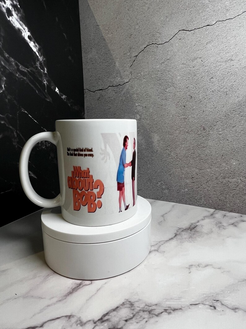 May include: White ceramic mug featuring the text "What About Bob?" in orange and an image of two men shaking hands. The mug also has the quote "He's a special kind of friend. The kind that drives you crazy."