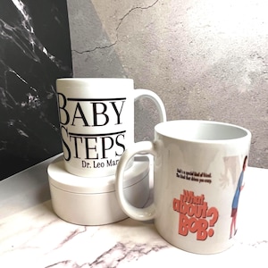 May include: Two white ceramic mugs. One mug has the words "BABY STEPS" printed in black, with "Dr. Leo Marvin" below. The other mug has the text "What about Bob?" in orange, with a cartoon image.