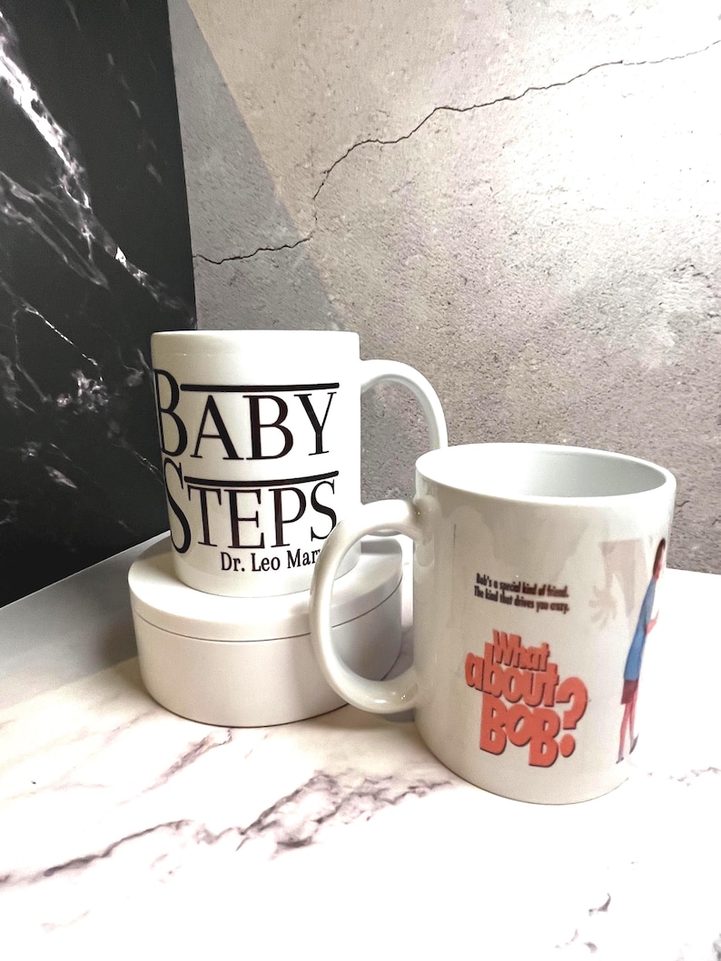 May include: Two white ceramic mugs. One mug has the words "BABY STEPS" printed in black, with "Dr. Leo Marvin" below. The other mug has the text "What about Bob?" in orange, with a cartoon image.