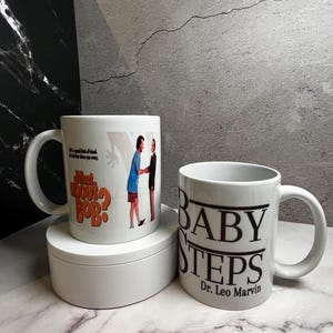 May include: Two white ceramic mugs. One mug features the text "What About Bob?" with an image of two men. The other mug has the text "BABY STEPS Dr. Leo Marvin" in black. Both mugs have a white handle.