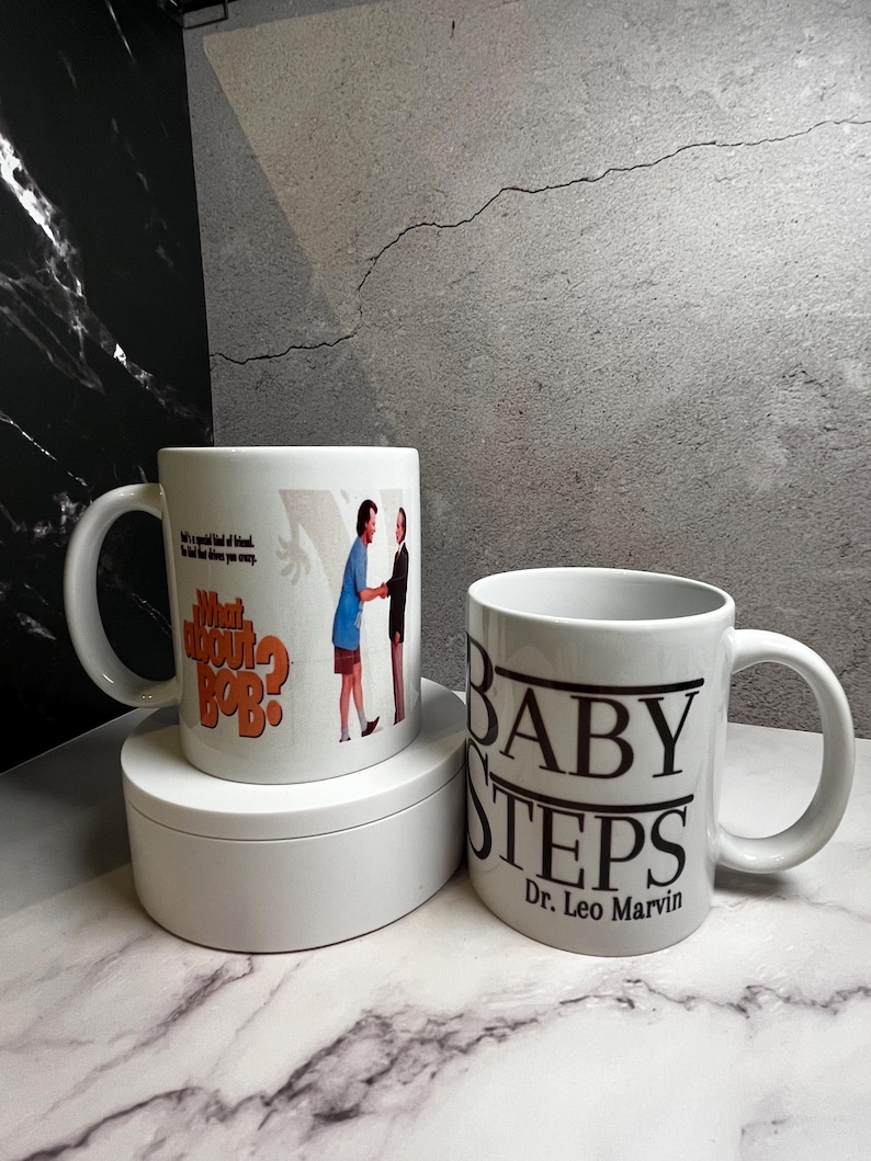 May include: Two white ceramic mugs. One mug features the text "What About Bob?" with an image of two men. The other mug has the text "BABY STEPS Dr. Leo Marvin" in black. Both mugs have a white handle.