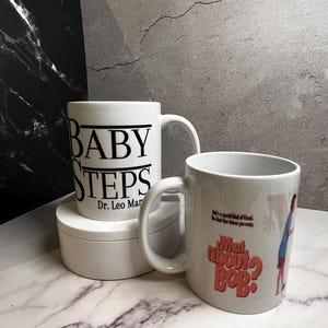 May include: Two white ceramic mugs. One mug has the words "BABY STEPS" and "Dr. Leo Marvin" printed in black. The other mug has the words "What About Bob?" and a cartoon image printed on it.