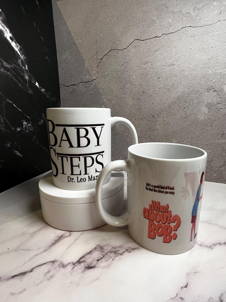 May include: Two white ceramic mugs. One mug has the words "BABY STEPS" and "Dr. Leo Marvin" printed in black. The other mug has the words "What About Bob?" and a cartoon image printed on it.