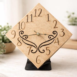 May include: A square wooden clock with engraved numerals and decorative flourishes. The clock face is a light tan color, with black numerals and hands. The clock is mounted on a black base.