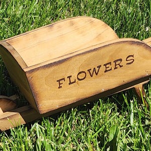 May include: A rustic wooden wheelbarrow planter with the word "FLOWERS" in dark brown lettering. The wheelbarrow is made of light-colored wood with darker brown trim and is set on a bed of green grass.