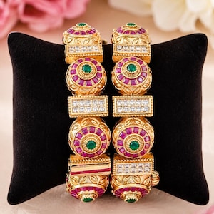May include: A pair of gold-toned bangles featuring alternating circular and rectangular segments. The circular sections are set with vibrant pink stones and a central green stone, while the rectangular segments are encrusted with clear white stones.
