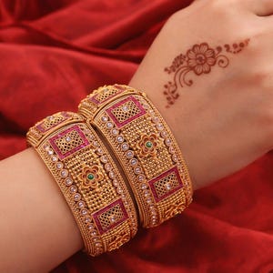 May include: Two gold-colored bangles with intricate designs, featuring red and green accents. The bangles are adorned with small, sparkling embellishments. The jewelry is displayed on a red fabric background.