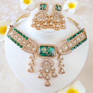 May include: A gold-toned necklace and matching earrings set. The necklace features a large rectangular green stone pendant with smaller green stones and clear crystals. The earrings have square green stones and dangling crystal accents. The jewelry is displayed on a white stand.