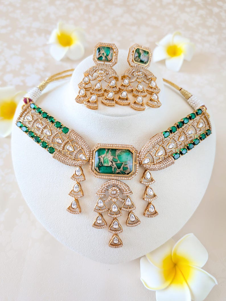 May include: A gold-toned necklace and matching earrings set. The necklace features a large rectangular green stone pendant with smaller green stones and clear crystals. The earrings have square green stones and dangling crystal accents. The jewelry is displayed on a white stand.