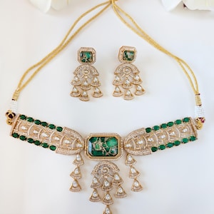 May include: A gold-toned necklace and earring set featuring emerald green and clear gemstones. The necklace has a central rectangular green stone and dangling teardrop-shaped accents. The earrings mirror the necklace design, with matching green stones and teardrop dangles.