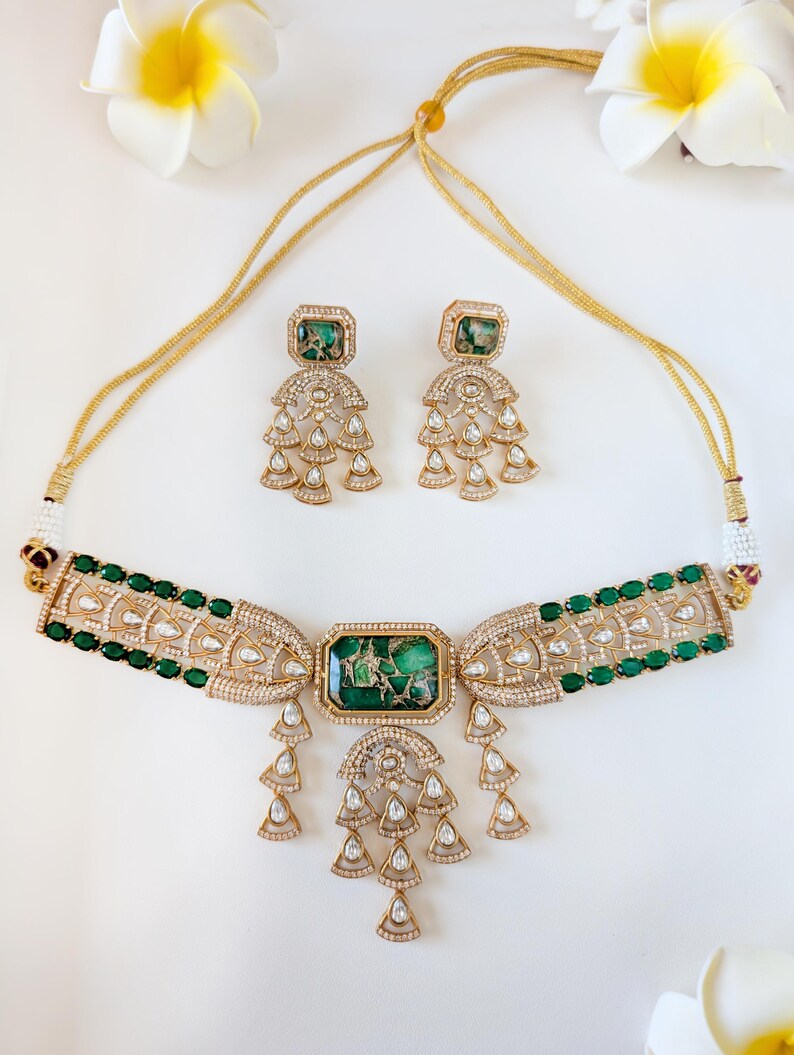 May include: A gold-toned necklace and earring set featuring emerald green and clear gemstones. The necklace has a central rectangular green stone and dangling teardrop-shaped accents. The earrings mirror the necklace design, with matching green stones and teardrop dangles.