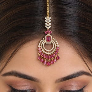 May include: A gold-toned maang tikka headpiece featuring a central rectangular red stone, surrounded by circular rows of clear white crystals, and finished with a fringe of dangling teardrop-shaped red stones, suspended from a delicate gold chain.