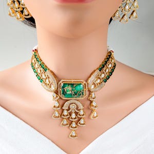 May include: A gold-toned necklace and matching earrings set. The necklace features a large, rectangular green stone pendant with intricate gold detailing and dangling teardrop-shaped accents. The earrings have a similar design.