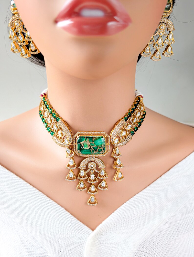 May include: A gold-toned necklace and matching earrings set. The necklace features a large, rectangular green stone pendant with intricate gold detailing and dangling teardrop-shaped accents. The earrings have a similar design.