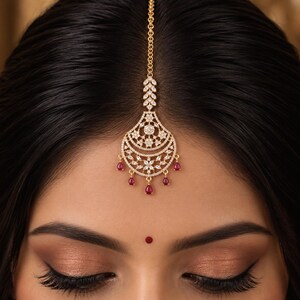 May include: A gold maang tikka headpiece featuring a central chain leading to a crescent-shaped pendant. The pendant is encrusted with clear white stones in a floral pattern and accented with five dangling teardrop-shaped red stones at the base.